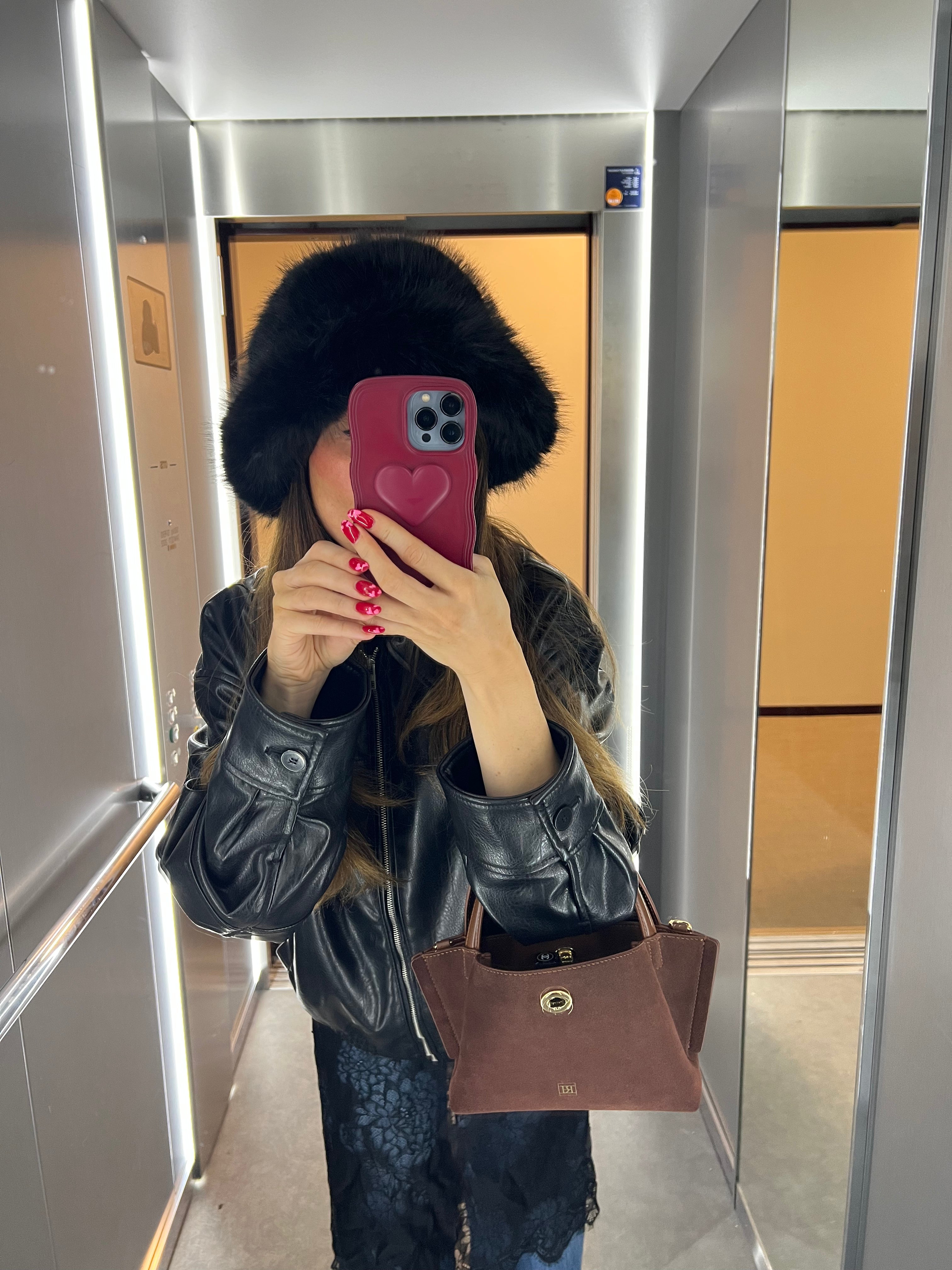 A fashionable girl taking a mirror selfie wearing a black leather jacket and fur hat, holding a phone with a pink case.