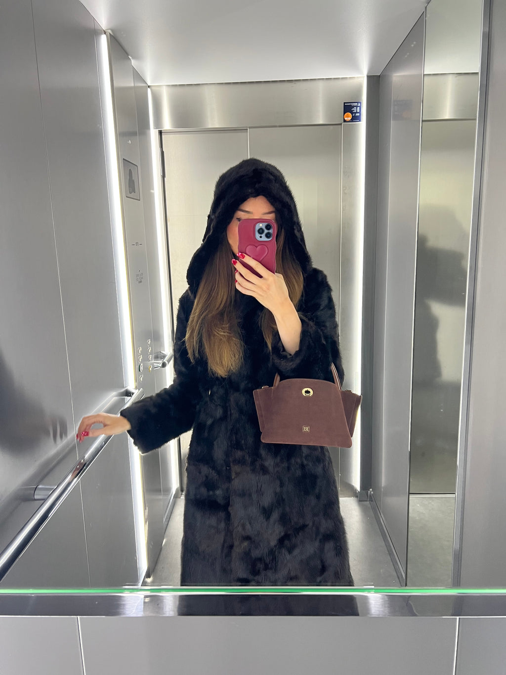 A girl taking a mirror selfie in an elevator wearing a dark coat and holding a brown handbag.