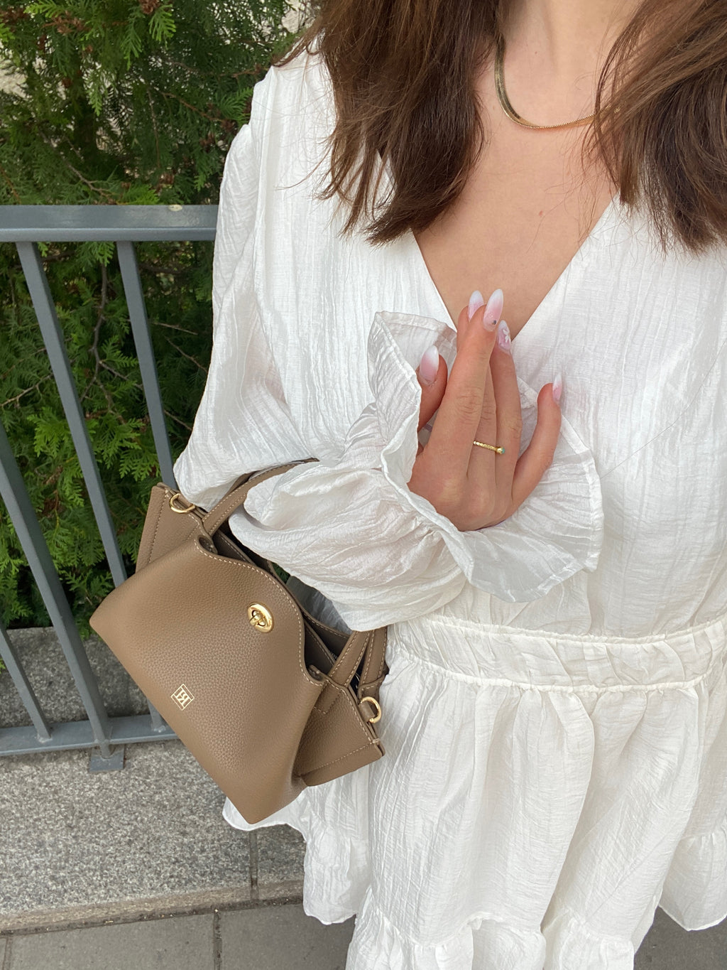 A girl wearing a white dress with a brown handbag in an outdoor background.