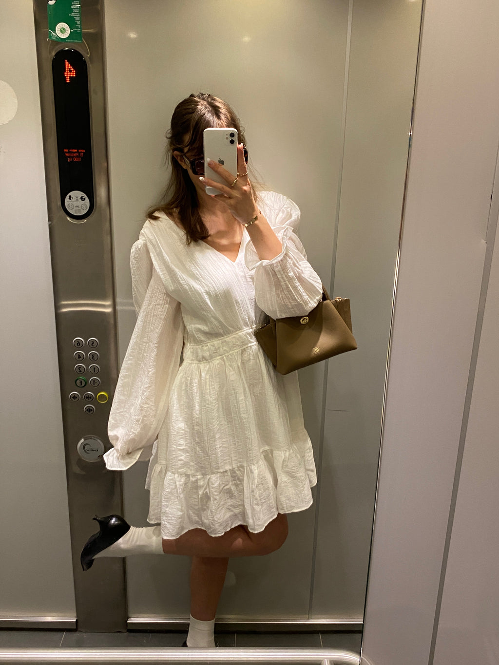 A girl in a white dress with a mini bag taking a selfie in an elevator.