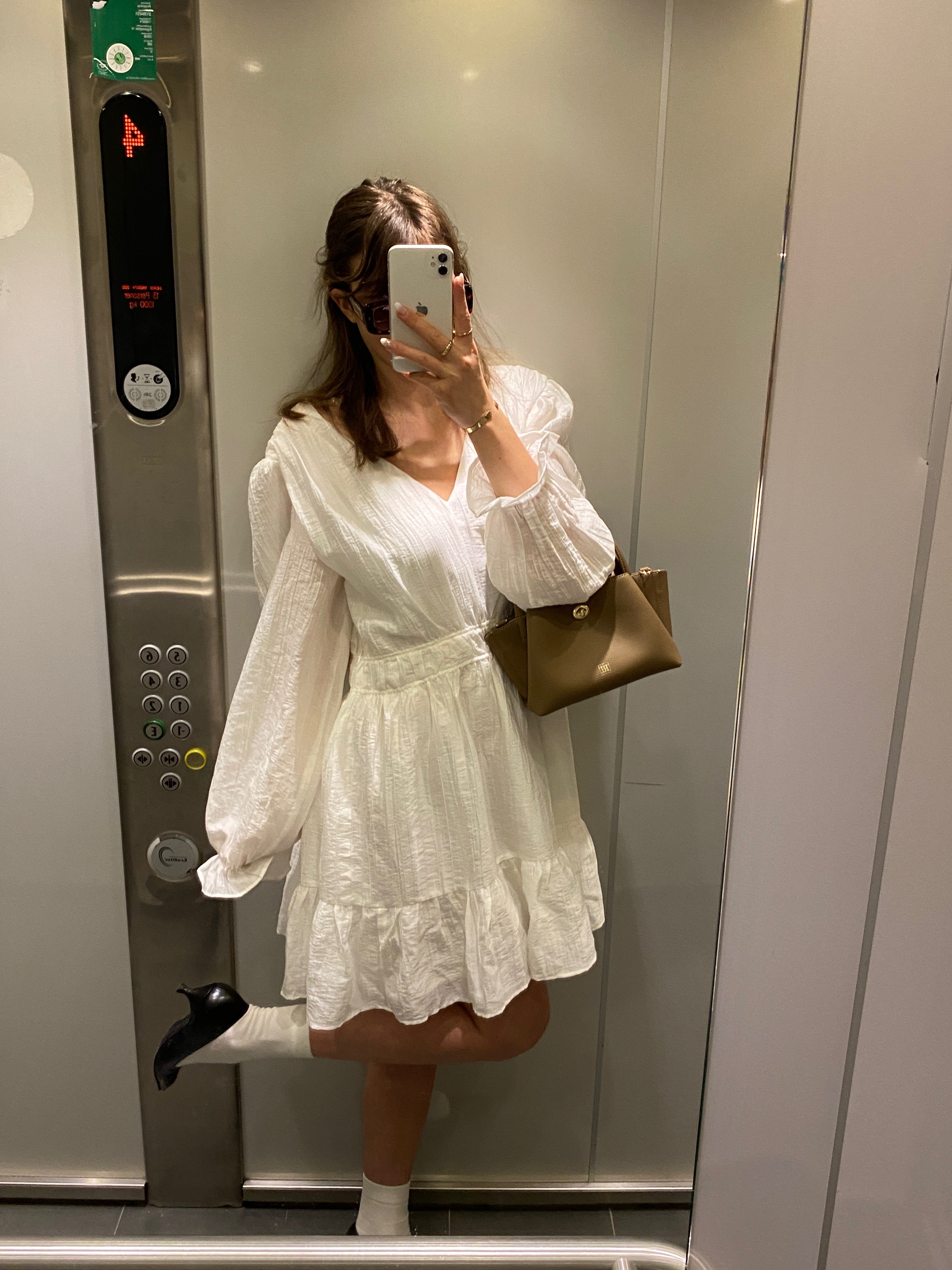A girl in a white dress with a mini bag taking a selfie in an elevator.